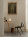 A black Ready Dining Chair stands beside a round wooden table. On the table is a lit Hashira Table Lamp, a book, and vases.