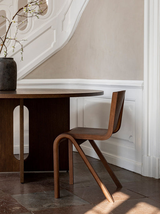 The oak Ready Dining Chair on a stone floor next to a dark table, with a white panelled wall behind.