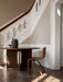 The wooden Ready Dining Chair sits by a dark round table in a sunlit hallway with a grand white staircase in the background.