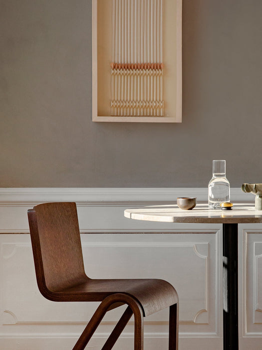 A dark wood Ready Dining Chair is placed next to a sunlit marble table. In the background is a grey wall with framed art.