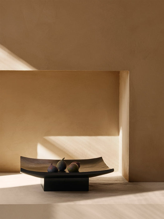The dark wood Relevé Platter with four figs, placed on a stone surface in a sunlit niche with strong shadows.