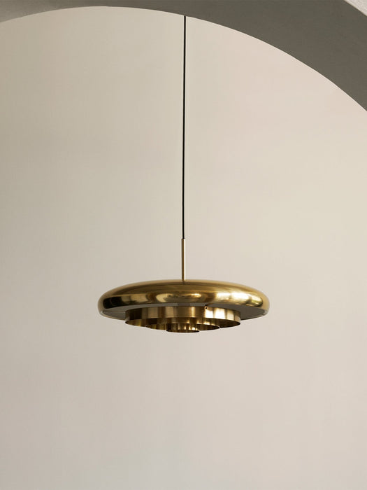 The Resonant Pendant, polished brass lamp with four concentric shades, hangs from a black cord against an off-white wall.
