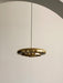 The Resonant Pendant, polished brass lamp with four concentric shades, hangs from a black cord against an off-white wall.