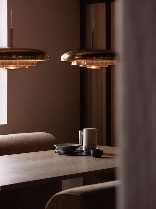Two polished brass Resonant Pendants illuminate a wooden dining table set with minimalist crockery in a warmly lit room.