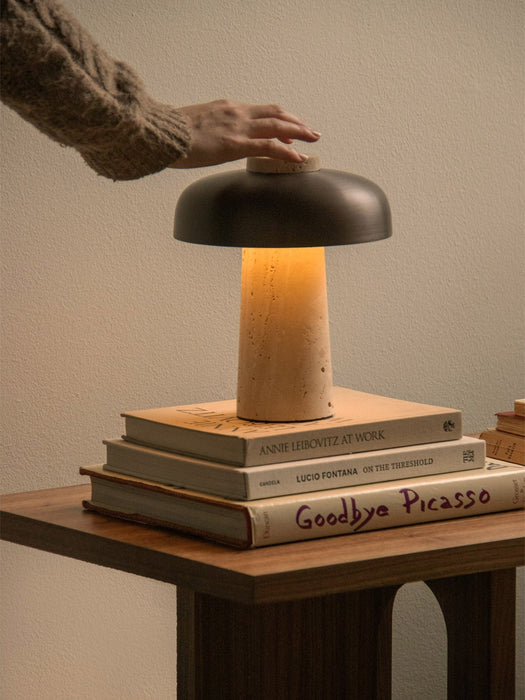 A hand adjusts the lit Reverse Table Lamp, Portable, which sits on a stack of art books on a wooden side table.
