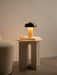 The lit Reverse Table Lamp, Portable sits on an Androgyne Side Table with a book and glass, beside a textured armchair.