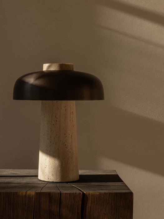 The Reverse Table Lamp, with its tactile travertine stone base and bronzed shade, sits on a dark wood block in soft light.