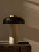 The Reverse Table Lamp, with its tactile travertine stone base and bronzed shade, sits on a dark wood block in soft light.