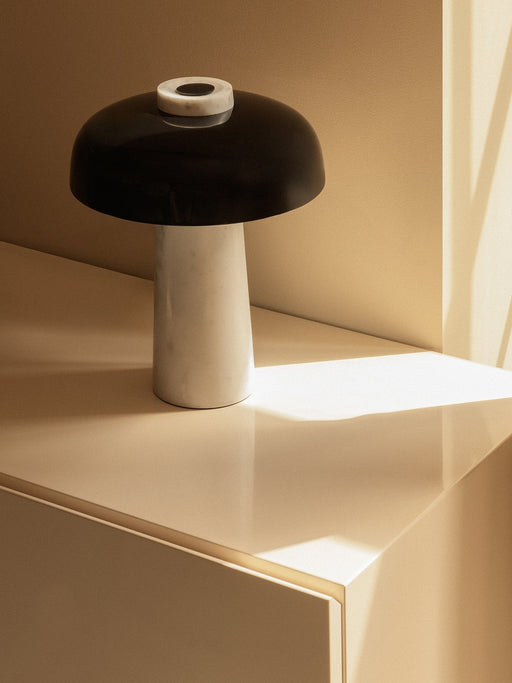 The Reverse Table Lamp with a white Carrara marble base and black shade, sitting on a cream cabinet in warm, direct sunlight.