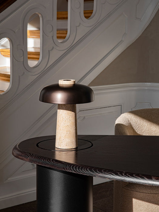 Reverse Table Lamp with a travertine base and bronzed shade, resting on a dark wood table in a sunlit room with a staircase.
