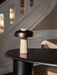 Reverse Table Lamp with a travertine base and bronzed shade, resting on a dark wood table in a sunlit room with a staircase.