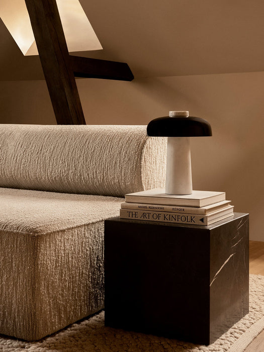 The Reverse Table Lamp on a stack of books atop a black Plinth Cubic table next to a cream Eave Modular Sofa in a cozy room.