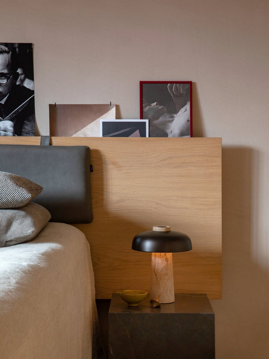 The Reverse Table Lamp with its travertine base and bronzed shade, sits lit on a dark marble plinth beside a modern bed.