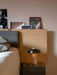 The Reverse Table Lamp with its travertine base and bronzed shade, sits lit on a dark marble plinth beside a modern bed.