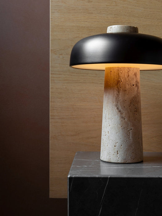 The Reverse Table Lamp with its travertine base and dark metal shade, illuminated on a dark marble plinth.