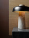 The Reverse Table Lamp with its travertine base and dark metal shade, illuminated on a dark marble plinth.