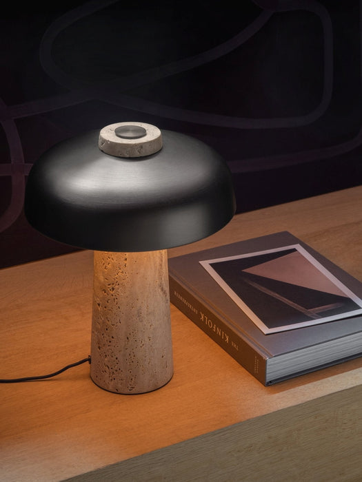 The Reverse Table Lamp, with a travertine stone base and black aluminium shade, sits lit on a wooden desk next to a book.
