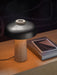 The Reverse Table Lamp, with a travertine stone base and black aluminium shade, sits lit on a wooden desk next to a book.