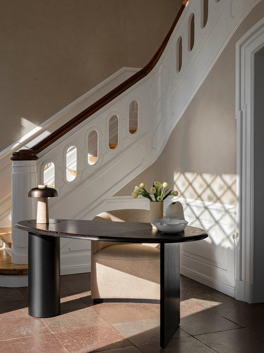 The Reverse Table Lamp sits on a dark oval desk in a sunlit entryway, with a grand white staircase in the background.