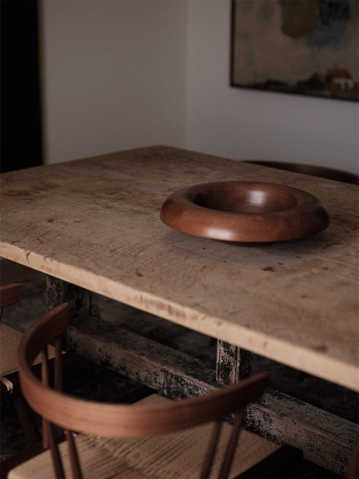 The Rond Bowl in dark wood sits on a rustic wooden table in a dimly lit room, with the back of a chair in the foreground.