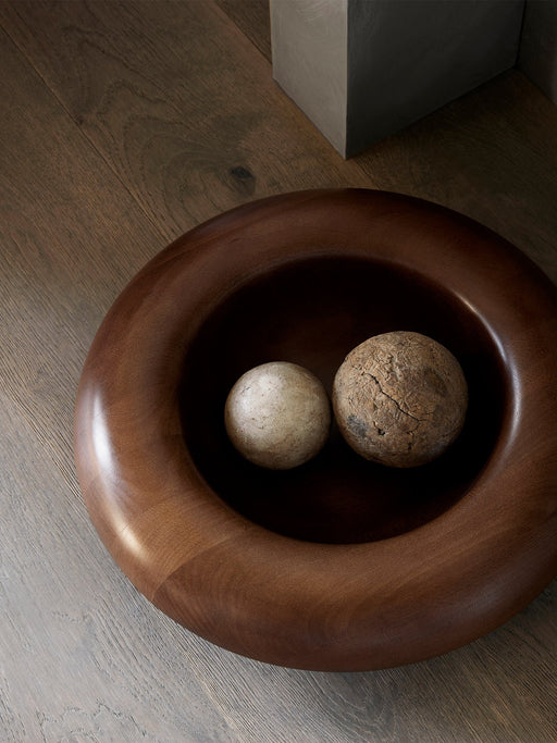 A large, dark brown wooden Rond Bowl on a wooden floor. Two decorative, textured stone balls sit inside.
