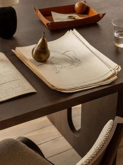 A close-up of a dark wooden desk with a Sentiment Paper Weight on a sketch, a Ready Dining Chair, and a Torso Table Lamp.