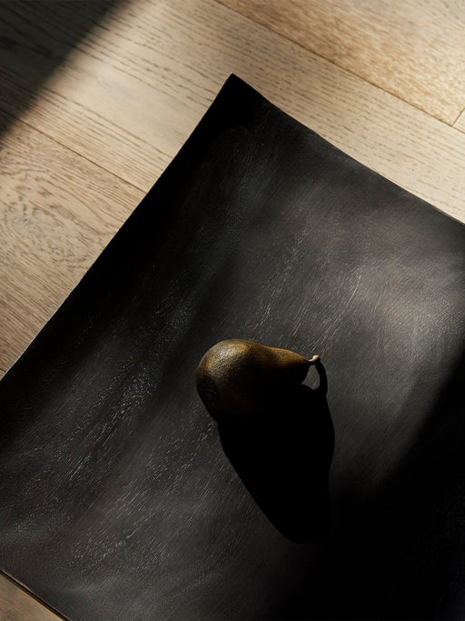 Top-down view of the bronze Sentiment Paper Weight on the black wood Releve Platter, with strong light and shadow on wood.