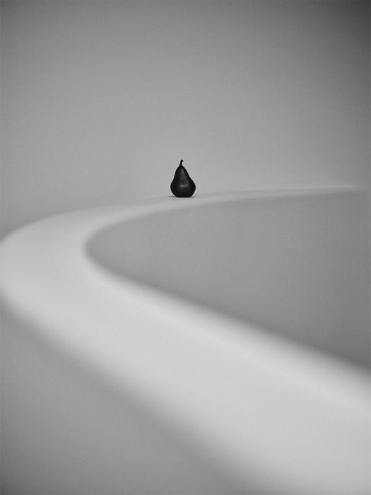 A minimalist black and white photograph of the Sentiment Paper Weight, a dark pear-shaped object, on a grey curved surface.
