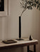 A black Strance Vase holds an olive branch on a marble table, by a brass Divot Tray and pear-shaped Sentiment Paper Weight.