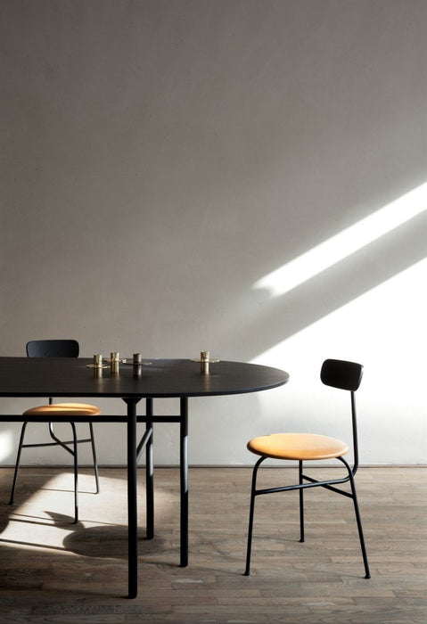 A black Snaregade Oval Table with two chairs is staged in a minimalist room with a wooden floor, lit by a diagonal sunbeam.