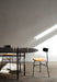 A black Snaregade Oval Table with two chairs is staged in a minimalist room with a wooden floor, lit by a diagonal sunbeam.
