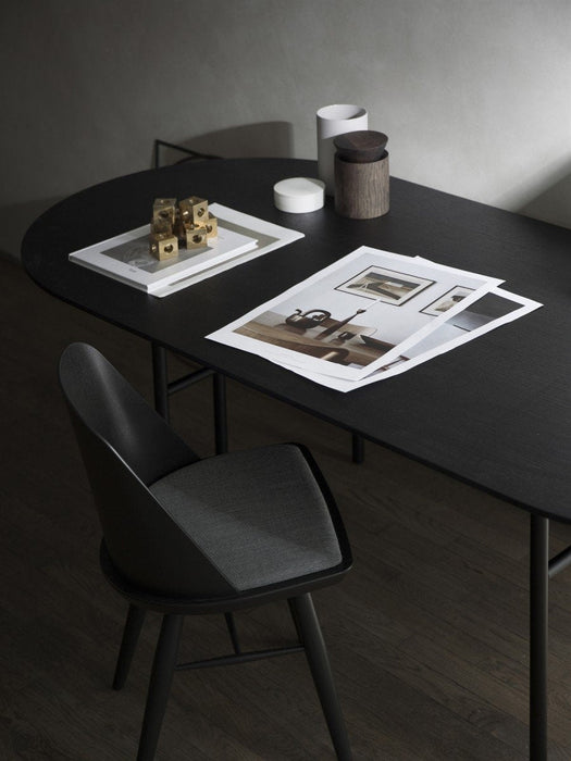 A black Snaregade Oval Table styled with brass cubes, papers and containers, viewed from above with a black dining chair.
