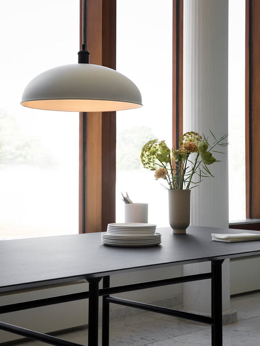 A Hubert Pendant hangs over a black Snaregade Dining Table set with plates and a Cyclades vase with flowers next to a window.