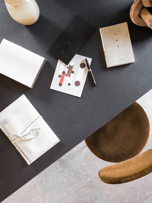 Overhead view of the Snaregade Dining Table styled with stationery, a pen, and books on a dark surface next to a brown chair.
