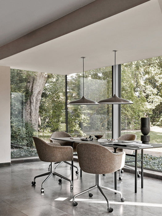 A black Snaregade Dining Table with brown wheeled chairs, set in a room with tiled floors and large windows facing a garden.