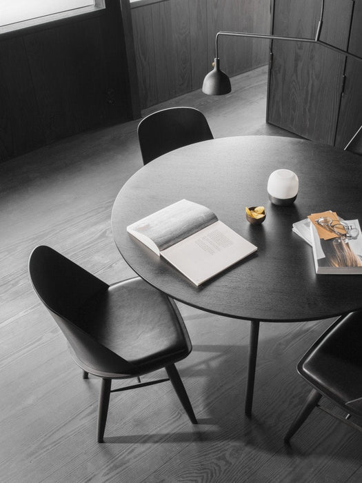 A black, round Snaregade Dining Table and Synnes Chairs styled with books and accessories in a dark, minimalist interior.