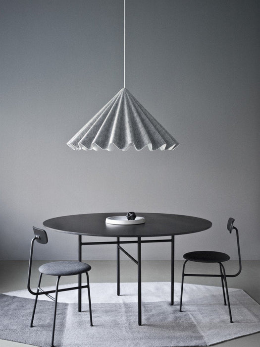 The black, round Snaregade Dining Table in a grey room with two chairs. A large, grey, pleated pendant lamp hangs above.