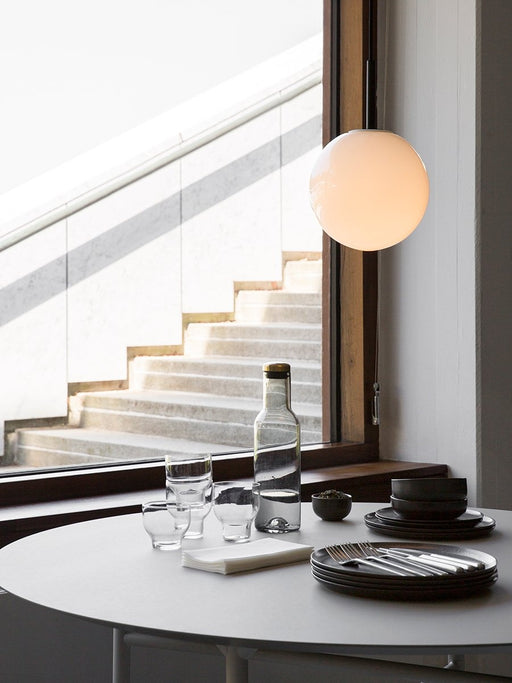 A view of the round Snaregade Dining Table set with dark plates and glassware under a glowing spherical pendant light.