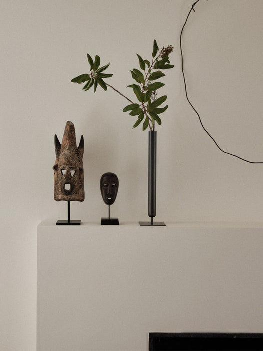 A tall, black Stance Vase holds a leafy branch, sitting on a white shelf alongside two carved wooden masks.