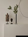 A tall, black Stance Vase holds a leafy branch, sitting on a white shelf alongside two carved wooden masks.