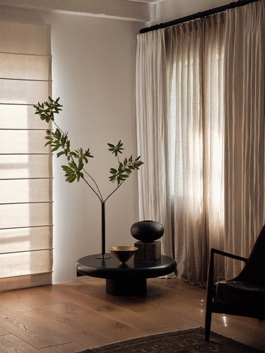 A black Stance Vase with a green branch, a gold Taper Bowl, and a black Surround Vase on a coffee table in a sunlit room.