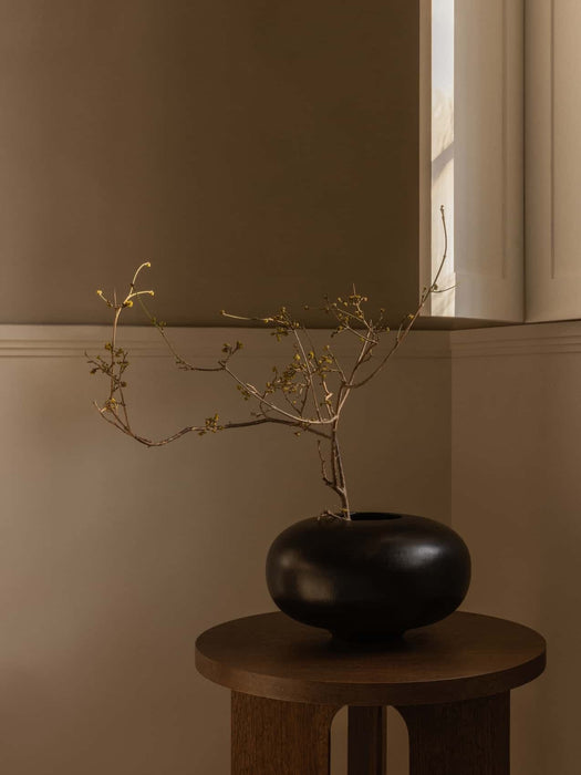 A black, rounded Surround Vase with a delicate, budding branch, sitting atop a dark wood Androgyne Side Table in a corner.