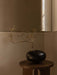 A black, rounded Surround Vase with a delicate, budding branch, sitting atop a dark wood Androgyne Side Table in a corner.