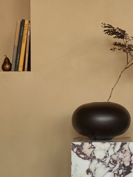 The dark brown Surround Vase holding a single branch, styled on a Calacatta Viola marble Plinth Pedestal by a wall shelf.