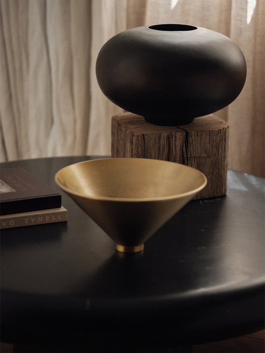 The golden Taper Bowl sits on a black table with books. In the background, the black Surround Vase rests on a wooden block.