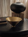 The golden Taper Bowl sits on a black table with books. In the background, the black Surround Vase rests on a wooden block.
