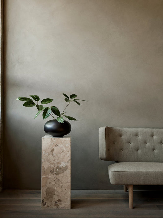 A black Surround Vase with green leaves rests on a tall marble Plinth Pedestal next to a grey tufted sofa in a modern room.