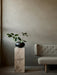 A black Surround Vase with green leaves rests on a tall marble Plinth Pedestal next to a grey tufted sofa in a modern room.