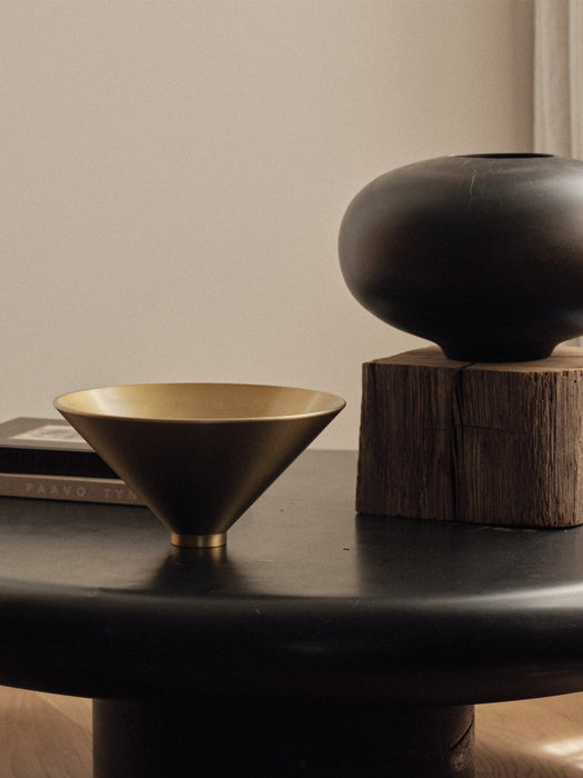 A close-up of a brass Taper Bowl and a dark, rounded Surround Vase. Both items are on a black table against a neutral wall.