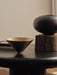 A close-up of a brass Taper Bowl and a dark, rounded Surround Vase. Both items are on a black table against a neutral wall.